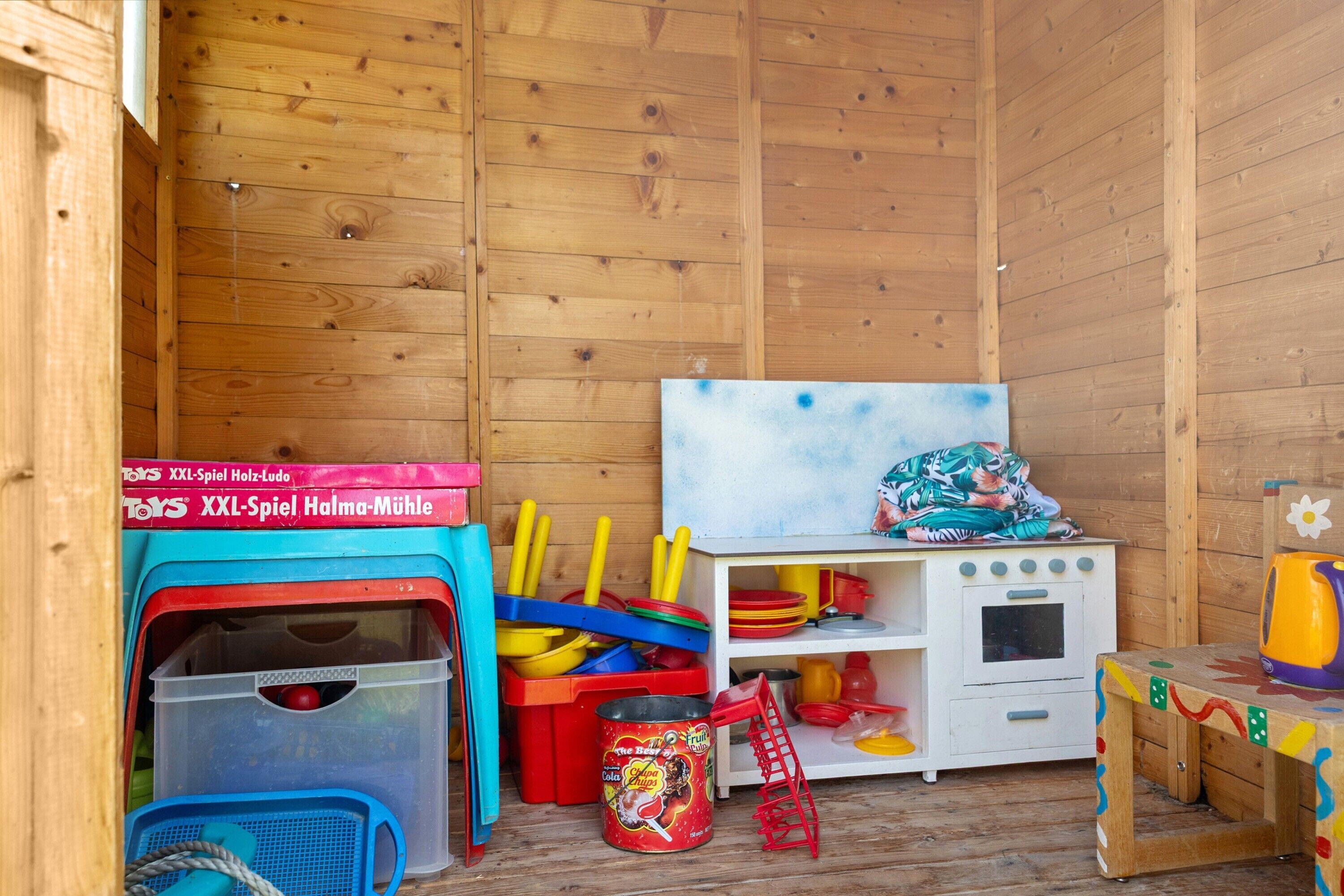 Um espaço de brincadeiras infantil em madeira, com brinquedos, uma cozinha de brinquedo e um casinha azul e vermelha.