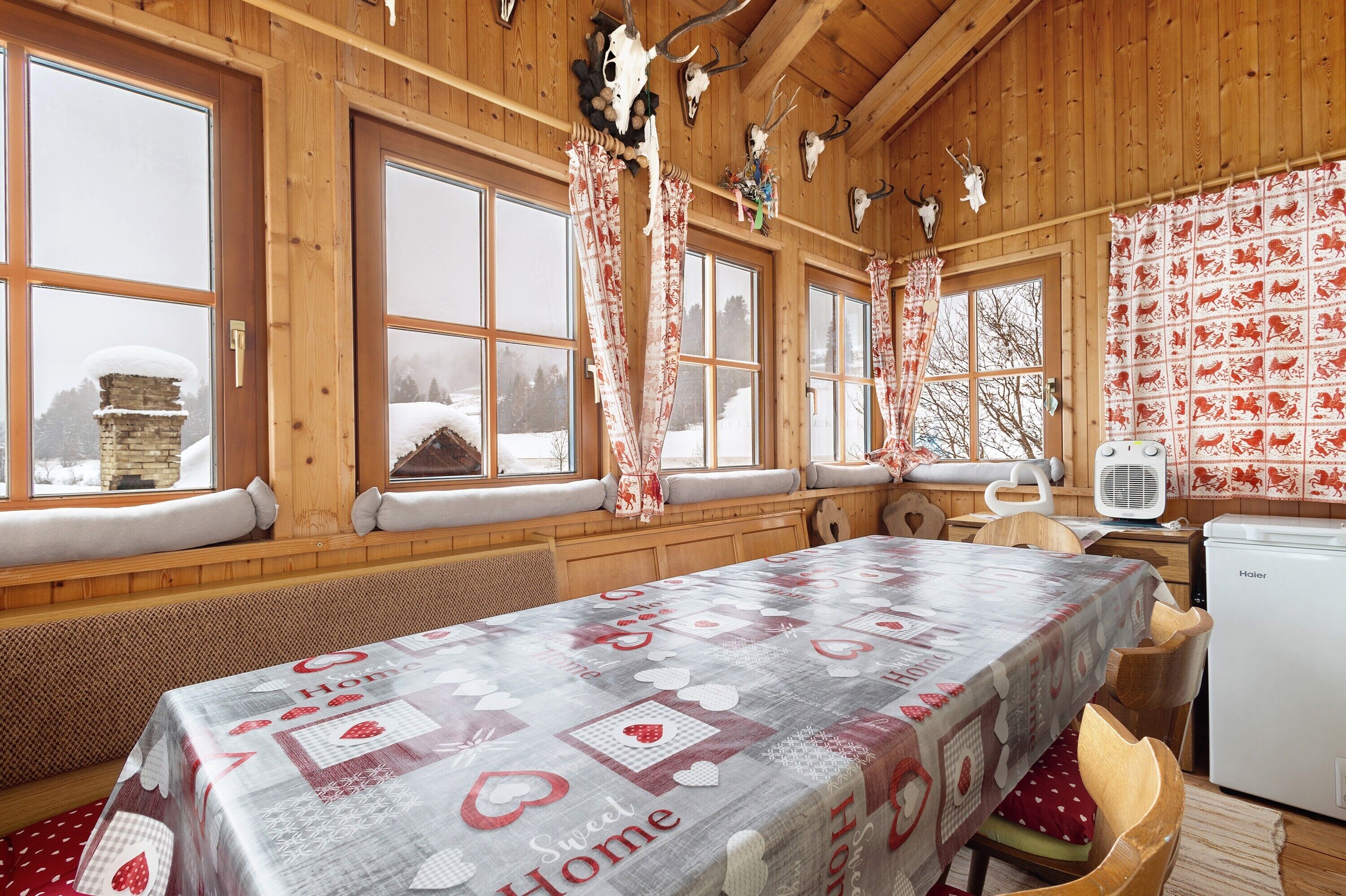 A cozy wooden cabin with large windows, a table with a heart-themed tablecloth, and a refrigerator.