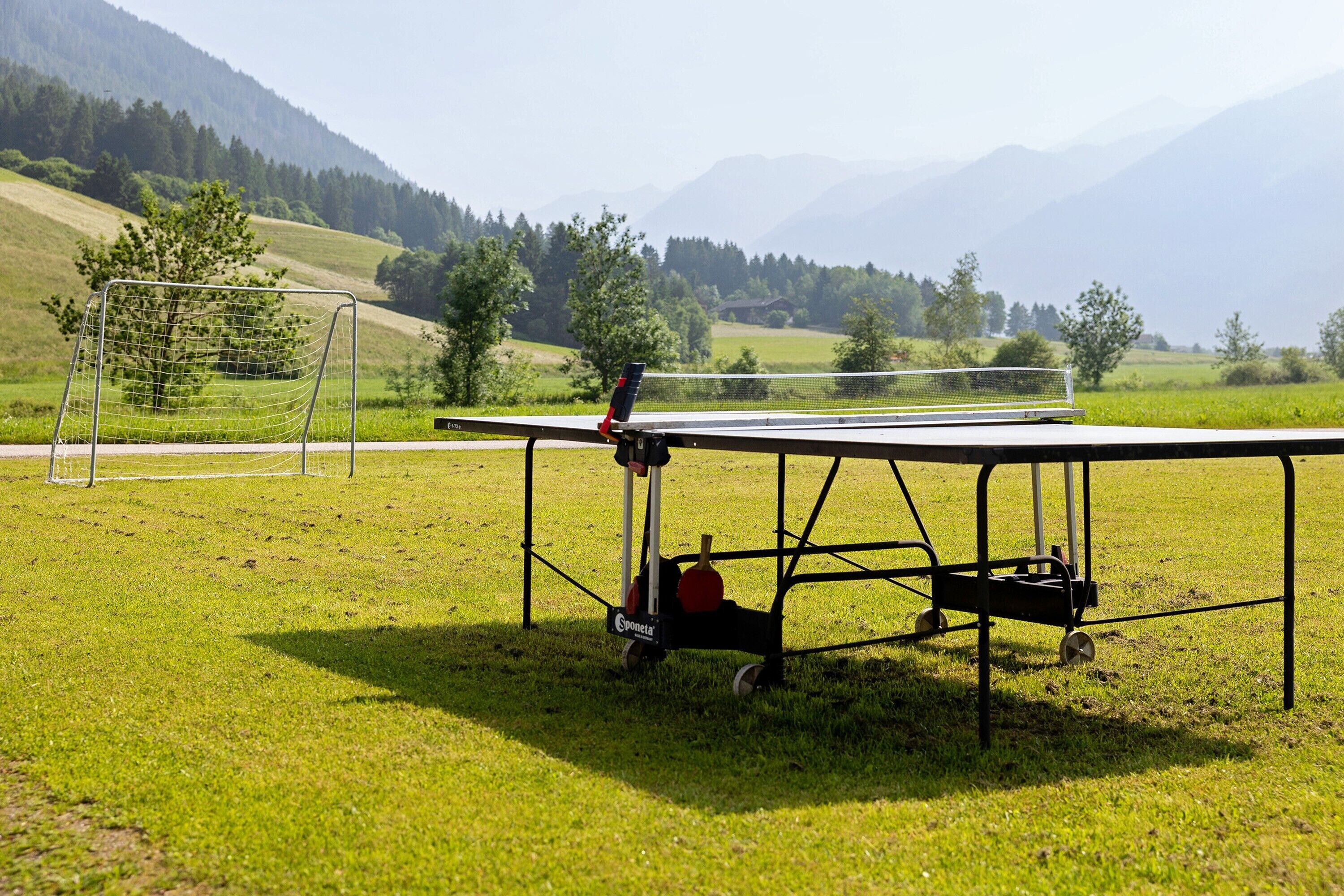 A ping pong table set up outdoors with a net, surrounded by a grassy field and mountains in the background.