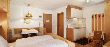 A compact hotel room with a bed, a small table, a kitchenette, and a seating area.