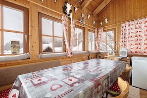 A cozy wooden cabin with large windows, a table with a heart-themed tablecloth, and a refrigerator.