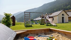 Un parque infantil con un tobogán y una estructura para escalar, un arenero con juguetes, y un edificio de techo inclinado en un área verde con montañas al fondo.