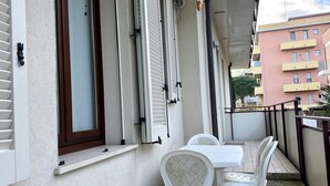 Een balkon met een witte tafel en stoelen, een raam met luiken en een leuning.