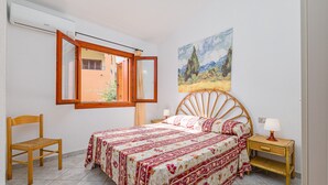 Een slaapkamer met een bed, een stoel, een nachtkastje, een lamp, een raam met een open luik, en een schilderij aan de muur.