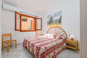 Un dormitorio con cama, una silla, una mesita de noche, una lámpara, una ventana con persiana abierta y un cuadro en la pared.