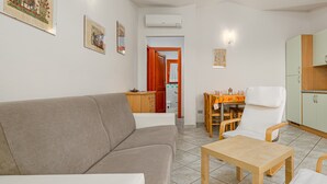 Een woonkamer met een bank, een houten tafel en een stoel. Er is een keuken met kastjes en een spoelbak.