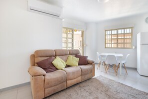 Una sala de estar con sofá, una mesa de comedor con sillas, un refrigerador y una ventana con persianas.