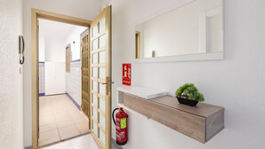 Un pasillo con una puerta de madera, un extintor, un espejo y una planta pequeña sobre una estantería.
