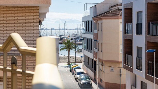 Vista de la calle con coches aparcados, un edificio con balcones y un puerto deportivo al fondo.
