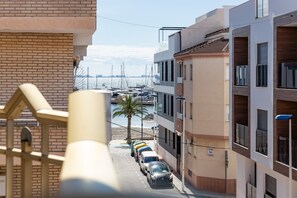 Vista de la calle con coches aparcados, un edificio con balcones y un puerto deportivo al fondo.