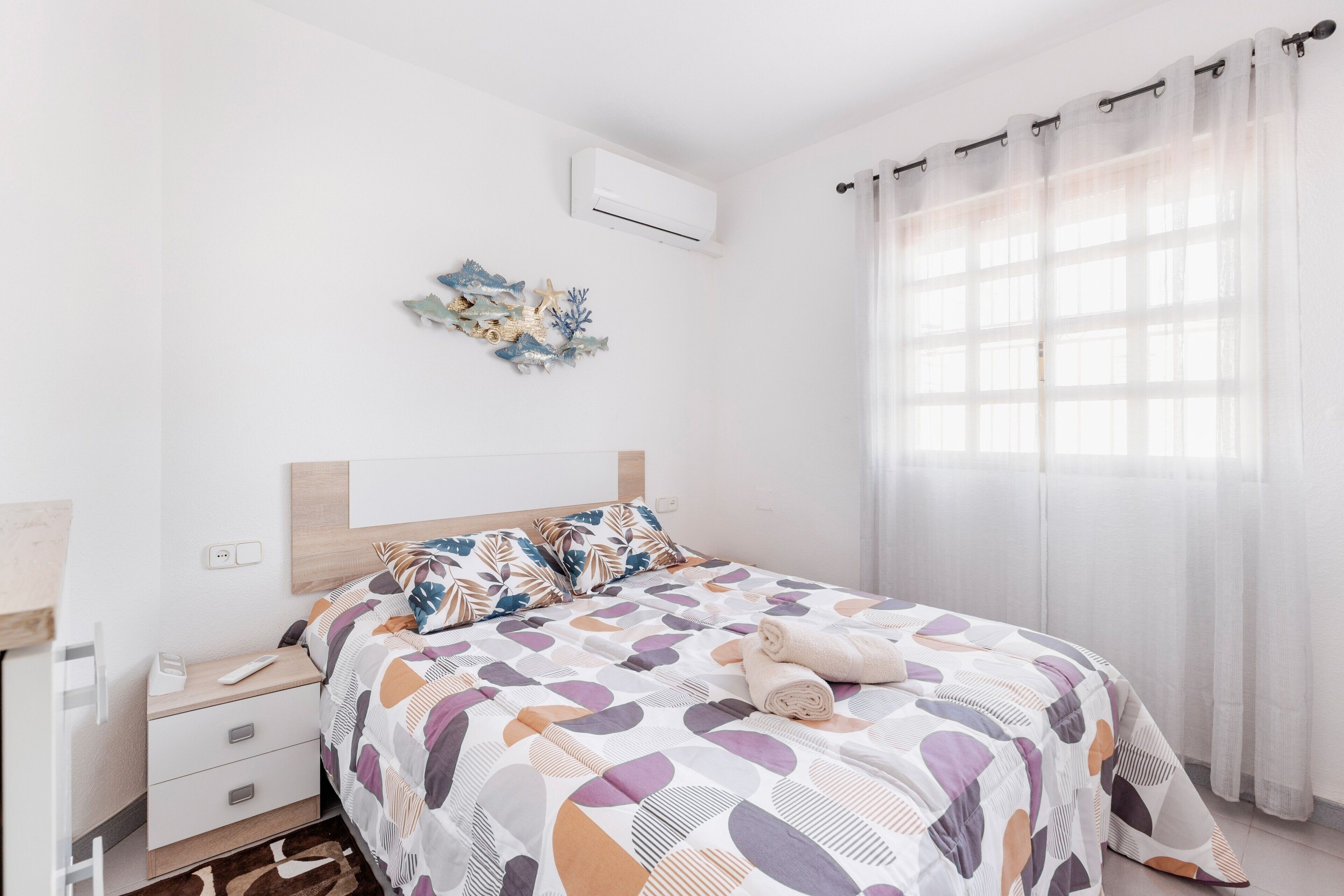 Un dormitorio con cama, mesita de noche, aire acondicionado de pared y ventana con cortinas traslúcidas.