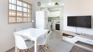 Una cocina moderna con televisor de pantalla plana, un comedor con mesa y sillas blancas, y vistas a través de una puerta abierta.
