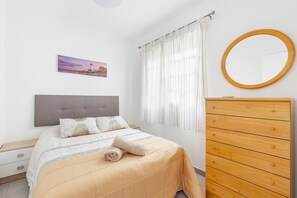 Un dormitorio con cama, cómoda de madera, espejo redondo, un cuadro y ventana con cortinas.