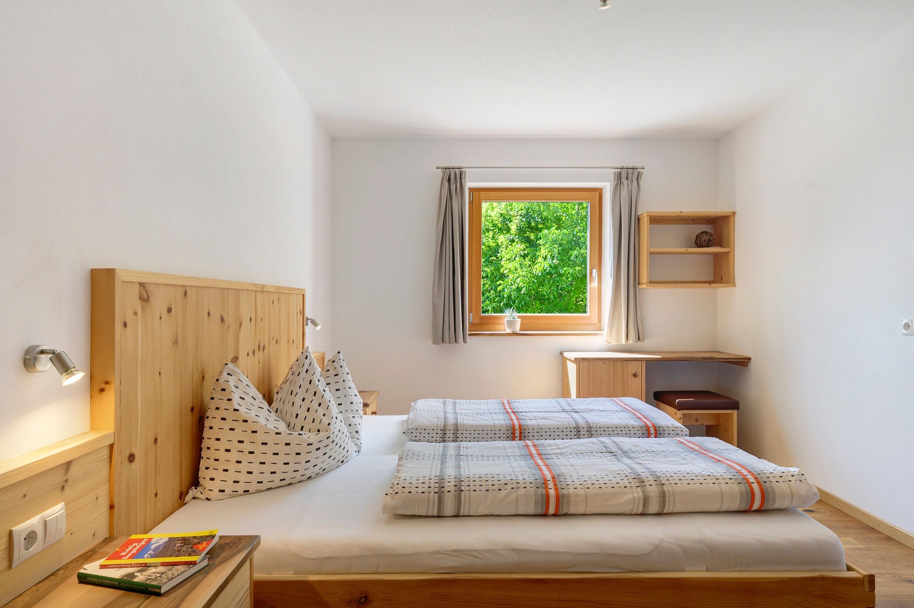 A bedroom with a wooden headboard, a bed with patterned bedding, a bedside table with a book, a window with curtains, and a small wooden shelf.