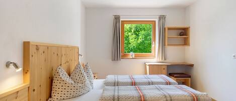 A bedroom with a wooden headboard, a bed with patterned bedding, a bedside table with a book, a window with curtains, and a small wooden shelf.