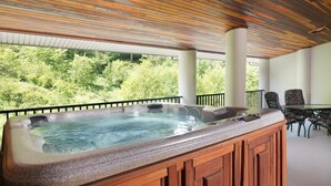 Una vasca idromassaggio con rivestimento in legno, un tavolo in vetro e sedie, su un balcone con vista sul verde.