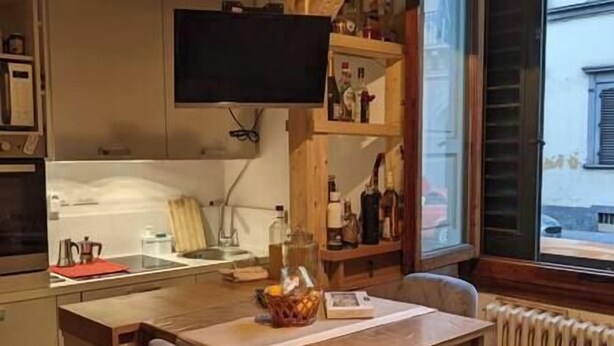 A kitchen with a wooden table, chairs, a TV mounted above the counter, and a window with a view of the street. - Small and cozy studio apartment in Florence (Florence)