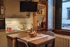 Een keuken met een houten tafel, stoelen, een tv boven de toonbank en een raam met uitzicht op de straat.