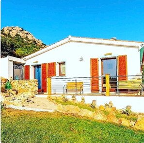 A white building with red shutters, a stone wall, and a grassy area in front. - Guest House 'Barbagia Plus' with Mountain View, Wi-Fi and Air Conditioning (Palau)