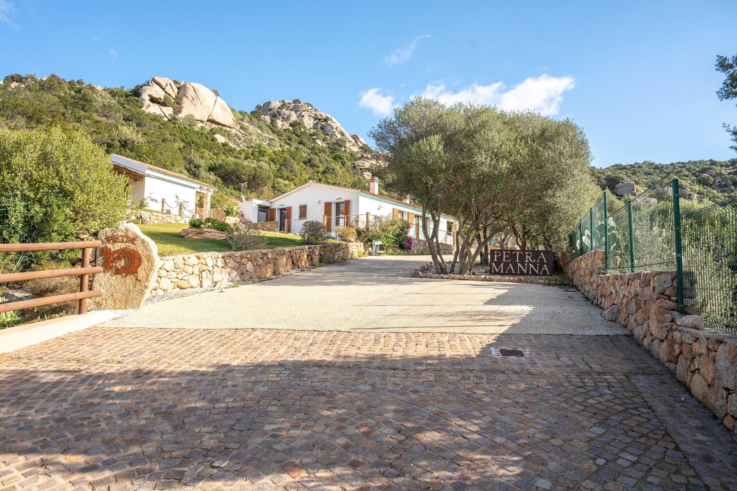 A paved pathway leading to a white building with a stone wall and a sign reading 'PETRA MANNA'.