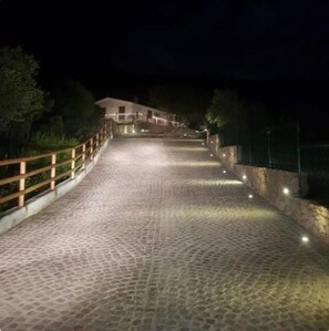 Un chemin pavé mène à un bâtiment bordé d’une clôture en bois d’un côté et d’un mur de pierre de l’autre, le tout éclairé la nuit.