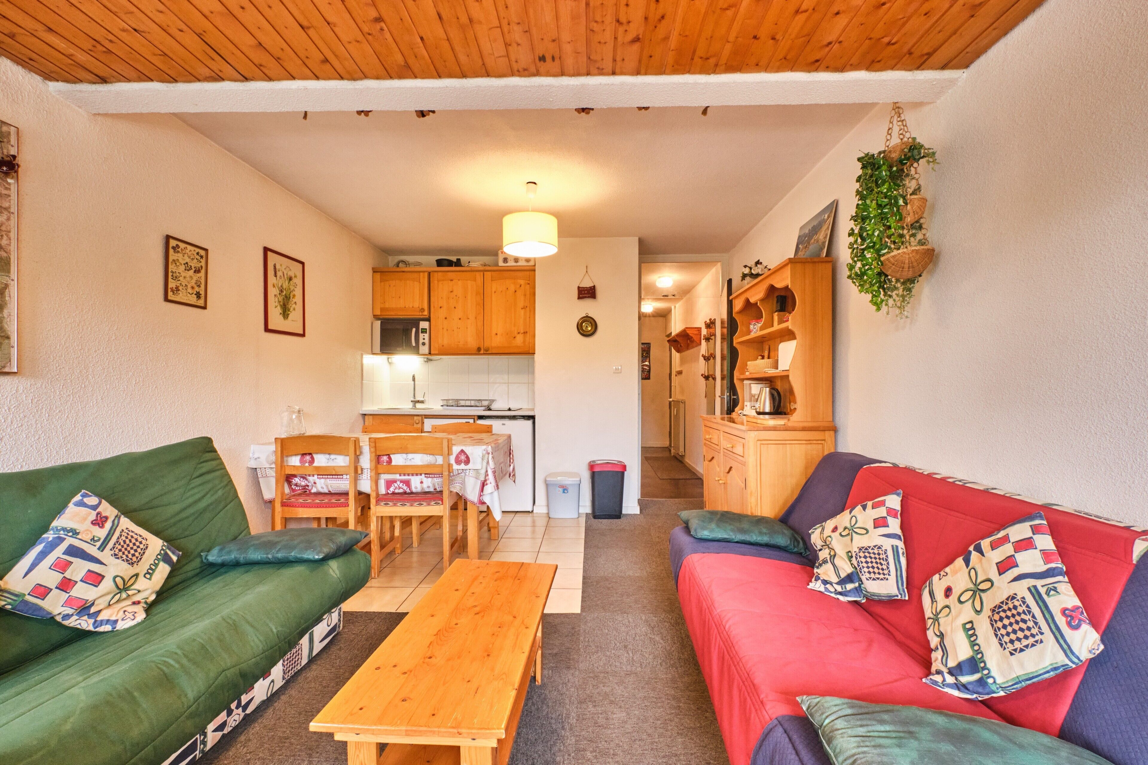 A cozy living room with a green sofa, a red sofa, a wooden table, and a kitchen in the background.