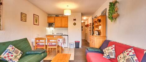 A cozy living room with a green sofa, a red sofa, a wooden table, and a kitchen in the background.