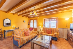 A living room with a sofa set, a glass coffee table, a wooden cabinet, and a television. - Casa del Tio Primitivo – Rural House with Mountain Views in Ávila (Navarredonda De Gredos)