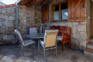 Un mur extérieur en pierre, un banc en bois, une table avec des chaises et une fenêtre avec des volets en bois.