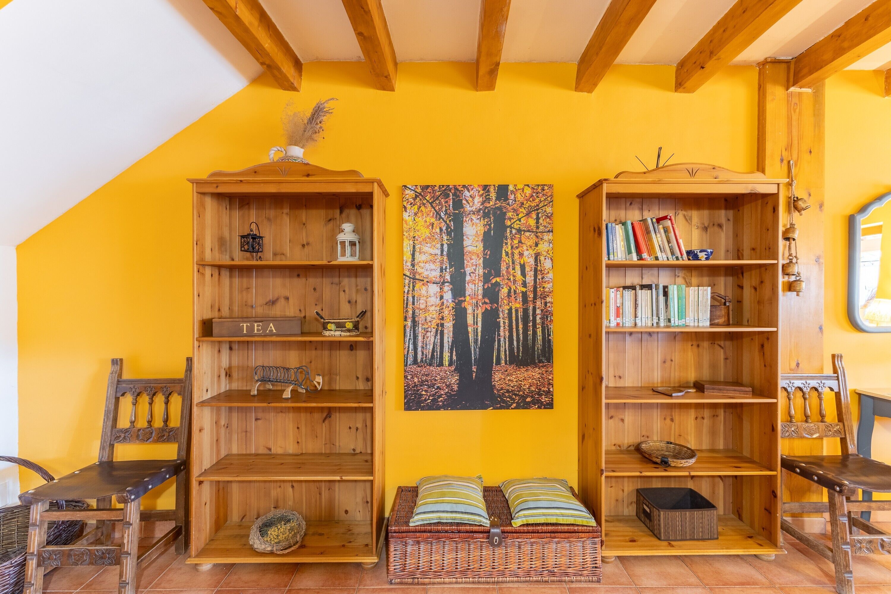 Una habitación con techo inclinado, estanterías de madera, un banco tejido y un cuadro colgado en la pared.