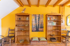 A room with a sloped ceiling, wooden bookshelves, a woven bench, and a wall-mounted painting. - Casa del Tio Primitivo – Rural House with Mountain Views in Ávila (Navarredonda De Gredos)