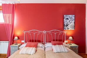Un lit double avec un cadre en métal blanc, deux tables de chevet avec des lampes, un mur rouge et une œuvre d’art florale au-dessus du lit.