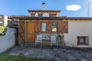 Un bâtiment rustique avec une façade en pierre, des volets en bois et un patio en pierre agrémenté de deux chaises blanches.
