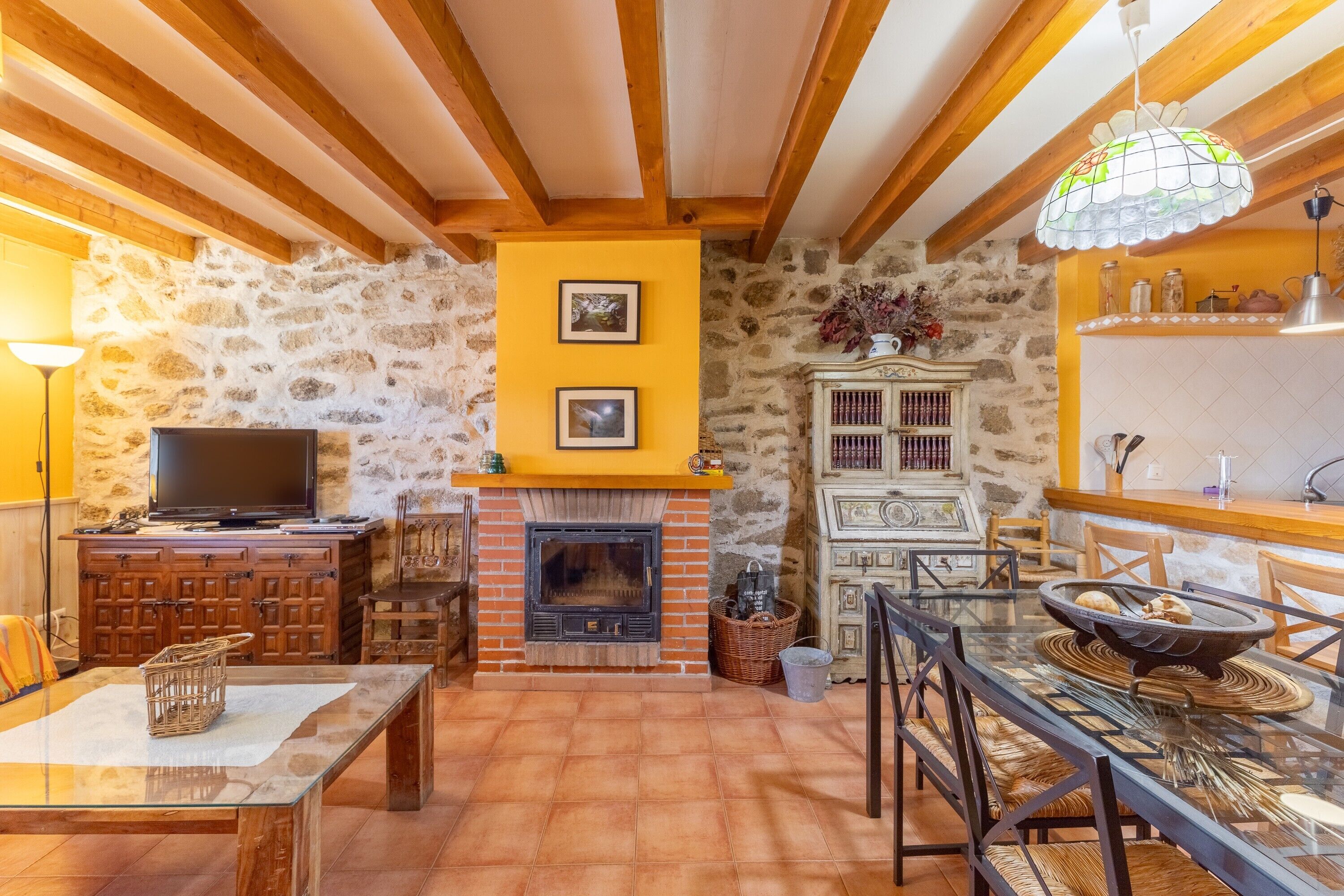Una sala de estar acogedora con chimenea, vigas de madera, paredes de piedra y una mesa de comedor de vidrio.
