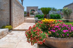 A stone pathway leading to a building with a stone wall, a large flower pot with various flowers, and a clear sky.