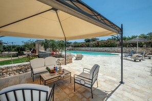 An outdoor patio area with a pool, stone flooring, outdoor furniture, and a pergola providing shade.