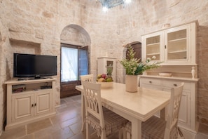 A stone-walled room with a television on a cabinet, a dining table with chairs, and a cabinet with glass doors.