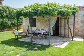 Un edificio in pietra con un portico coperto, un tavolo e sedie, e una pergola ricoperta di viti.