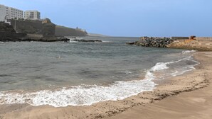 Escena costera con una playa arenosa, afloramientos rocosos y un edificio en un acantilado.