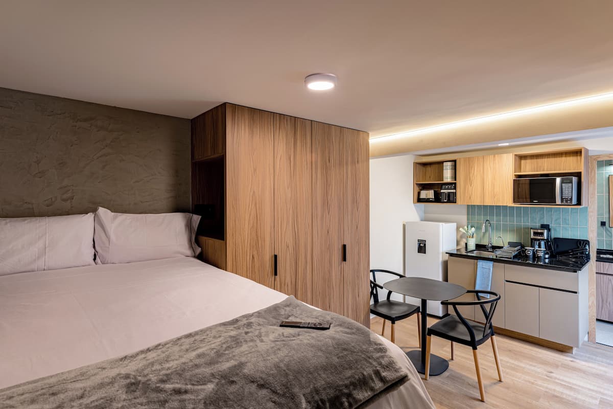 A bedroom with a large bed, a bedside table, and a small kitchen area with a microwave, sink, and cabinets.