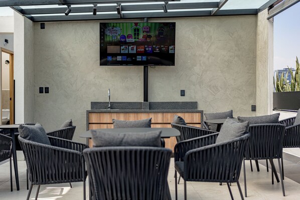 A modern outdoor lounge area with a flat-screen TV mounted on the wall, a bar counter, and several black wicker chairs. - Mint Roma Residences (Mexico City)