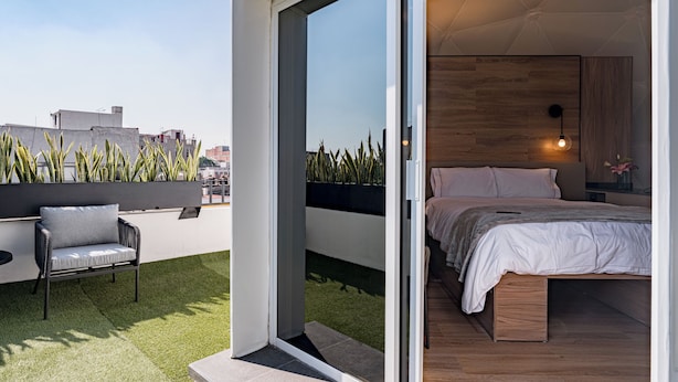 A modern bedroom with a large bed, a small seating area, and a view of the cityscape through a sliding door. - Mint Roma Residences (Mexico City)