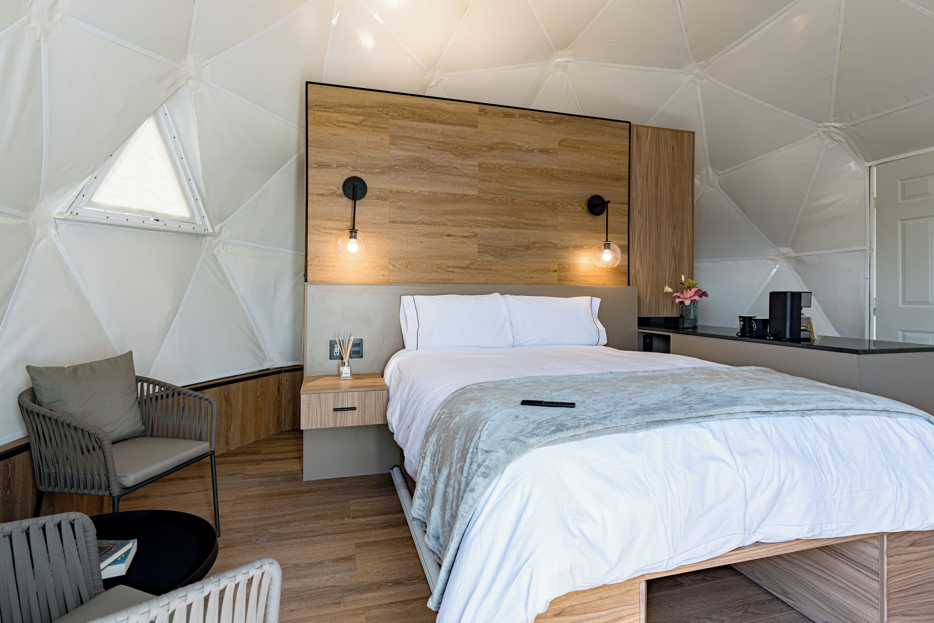 Un dormitorio con una cama grande, cabecera de madera, mesitas de noche, una silla y una ventana pequeña.