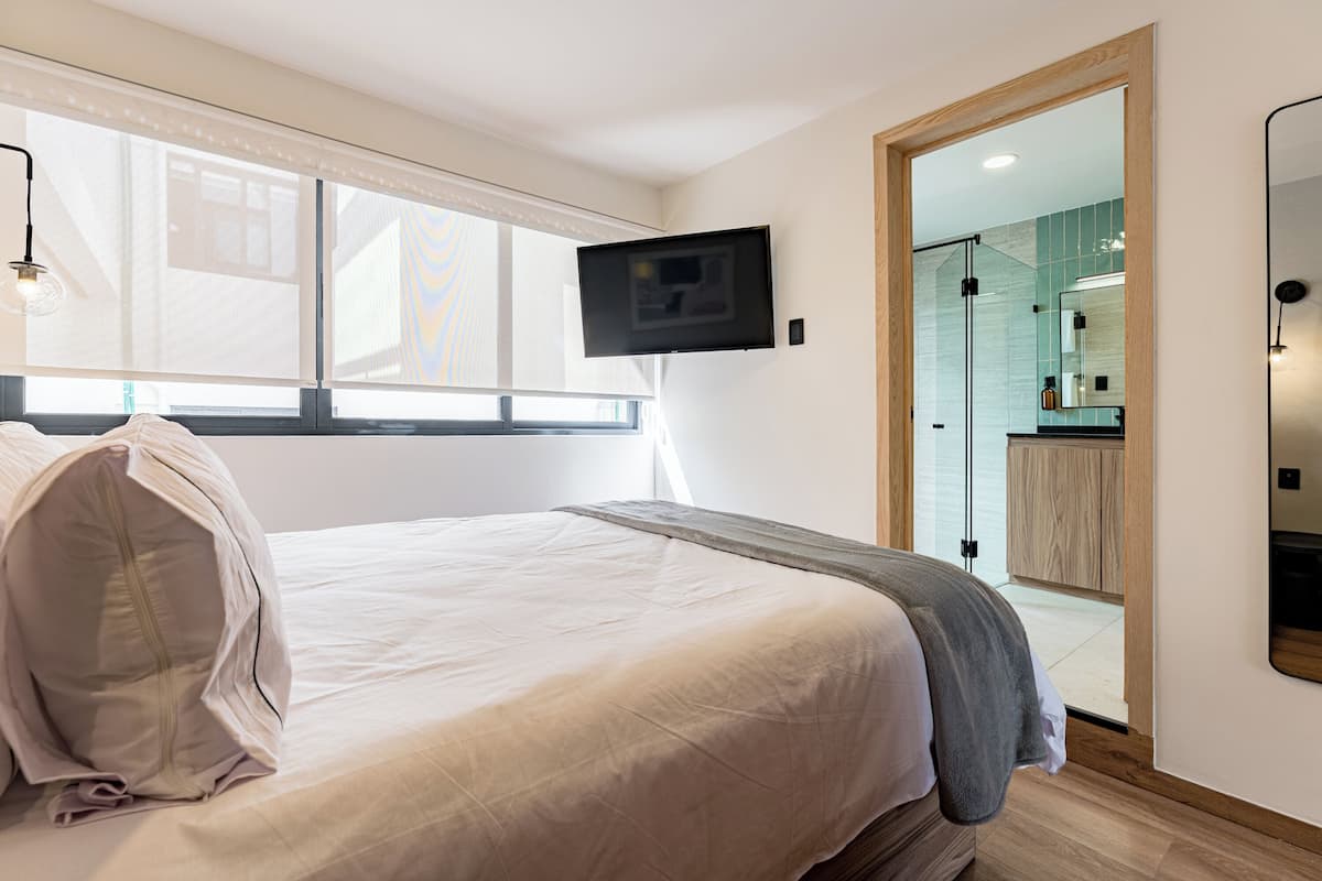 A modern hotel room with a large bed, a flat-screen TV mounted on the wall, and a bathroom visible through an open door.