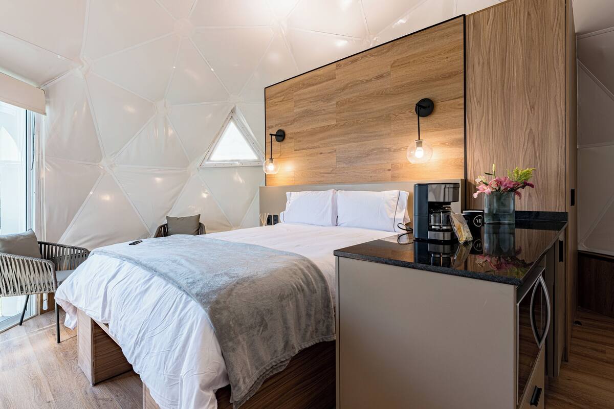 A modern bedroom with a large bed, a wooden headboard, a small kitchenette, and a geometric ceiling design.