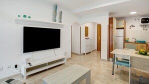 Ein modernes Wohnzimmer mit Flachbildfernseher, einer weißen Unterhaltungsecke, einem Essbereich mit Tisch und Stühlen sowie einer Küche mit Kühlschrank und Spüle.