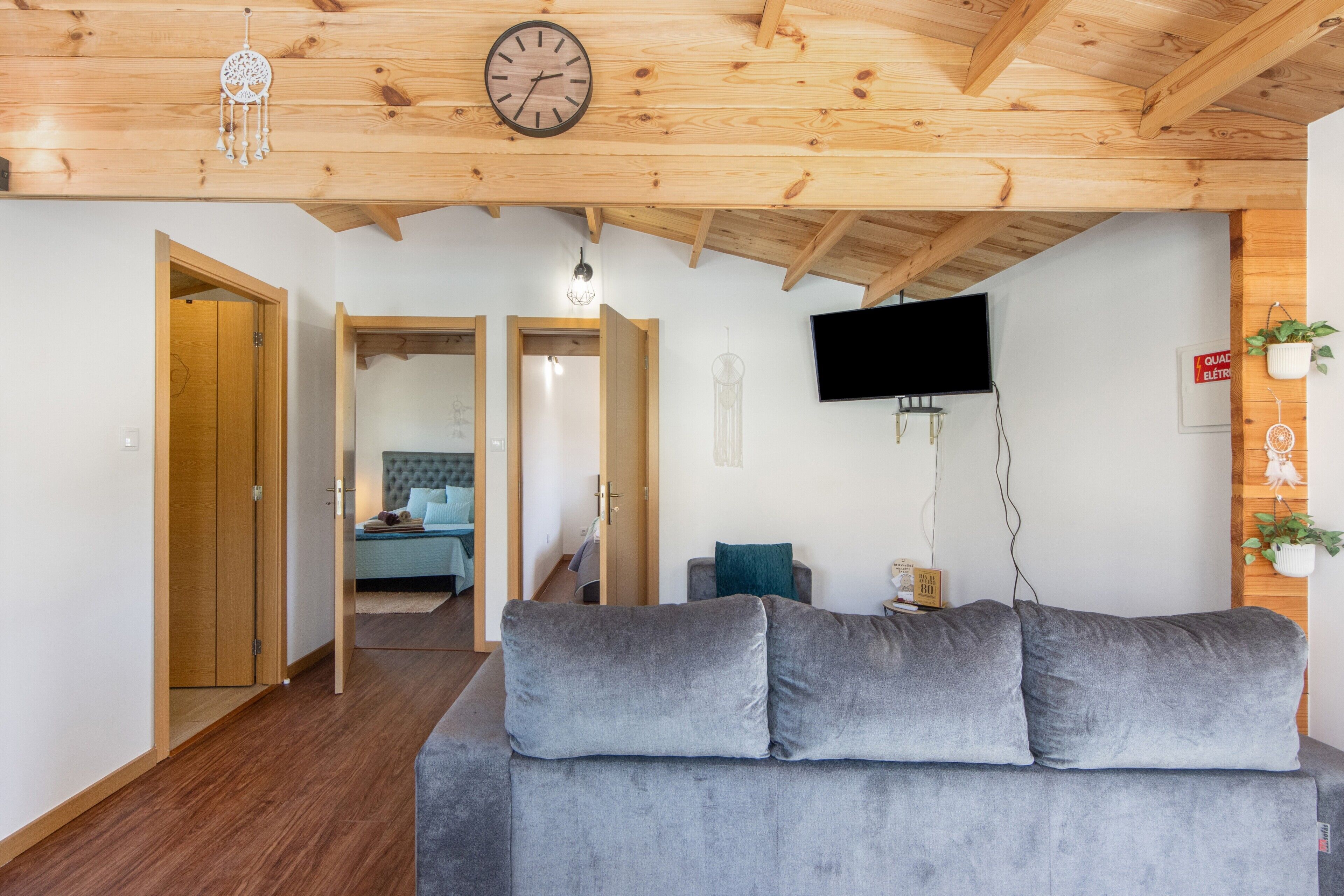 Una sala de estar con un sofá gris, vigas de madera en el techo, un televisor montado en la pared y una puerta que da a otra habitación.