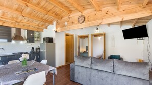 A modern kitchen with a dining area, a grey sofa, wooden ceiling beams, and a flat-screen TV mounted on the wall.
