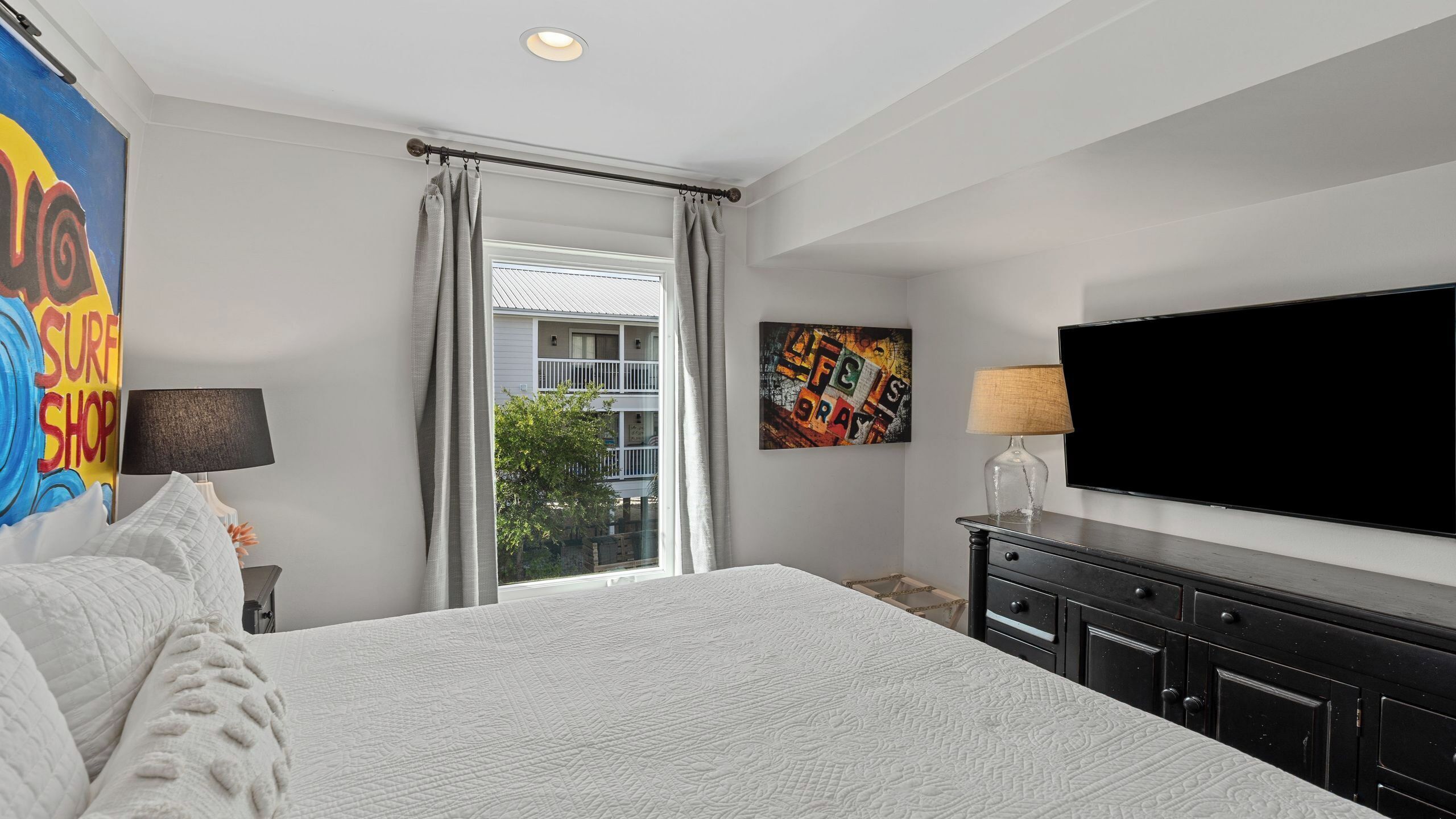 A bedroom with a large bed, a flat-screen TV, a dresser, a window with curtains, and a painting on the wall.