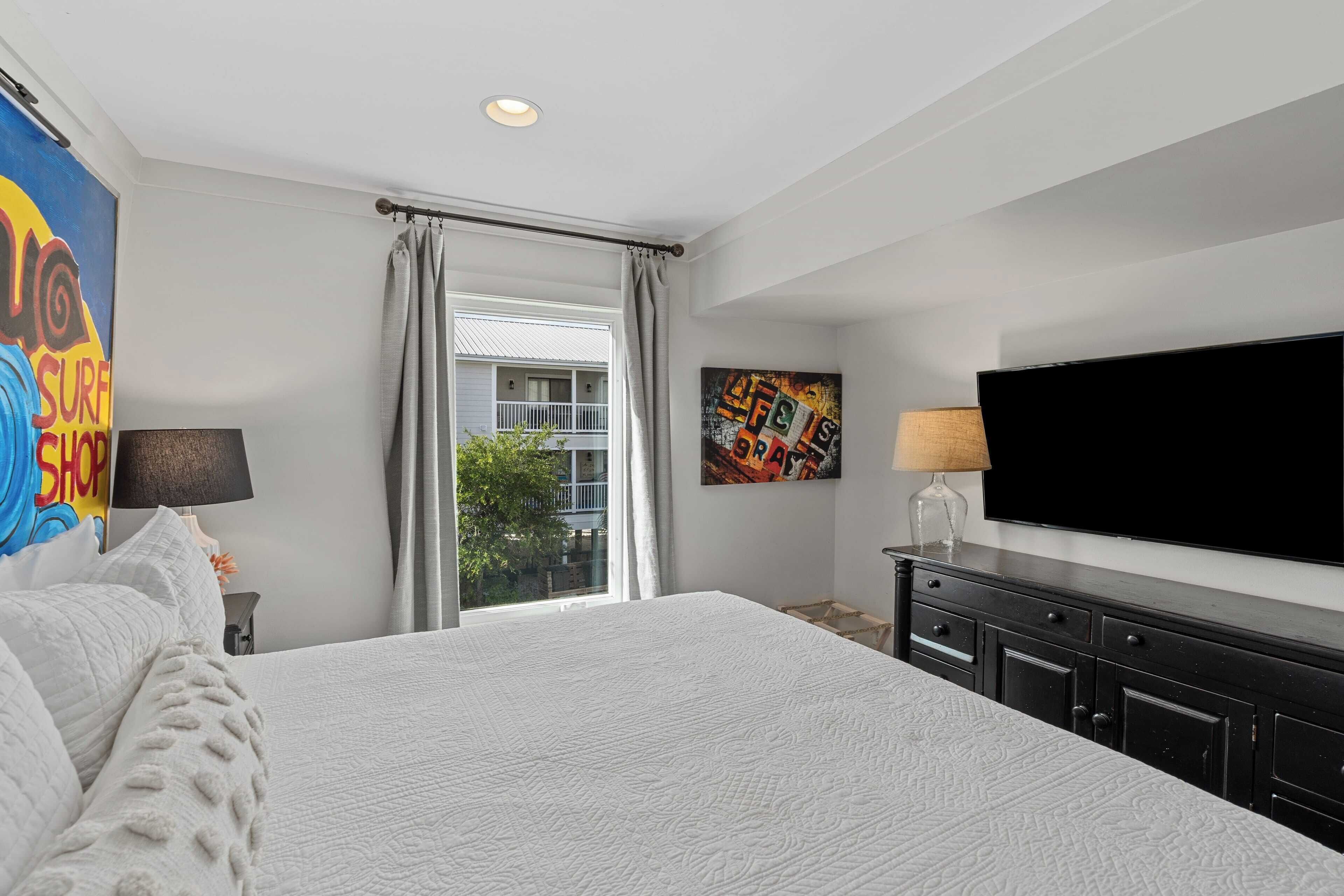 A bedroom with a large bed, a flat-screen TV, a window with curtains, and artwork on the walls.